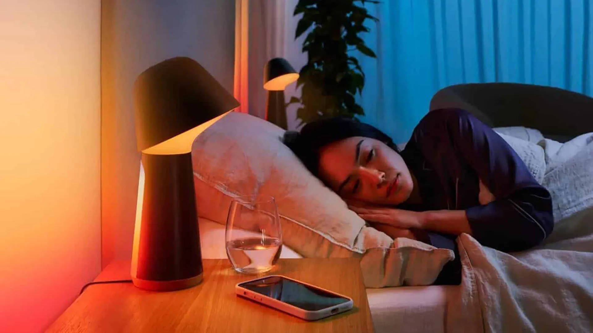 Người phụ nữ đang ngủ trên giường, cạnh đó là đèn ngủ thông minh và một chiếc smartphone, minh họa việc sử dụng điện thoại để tạo tiếng ồn trắng khi ngủ.