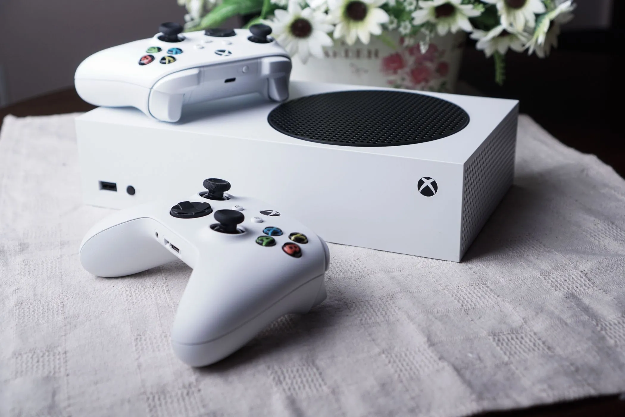 Máy chơi game Xbox Series S màu trắng đặt trên bàn cùng hai tay cầm và bình hoa, minh họa cho khả năng tối ưu game.