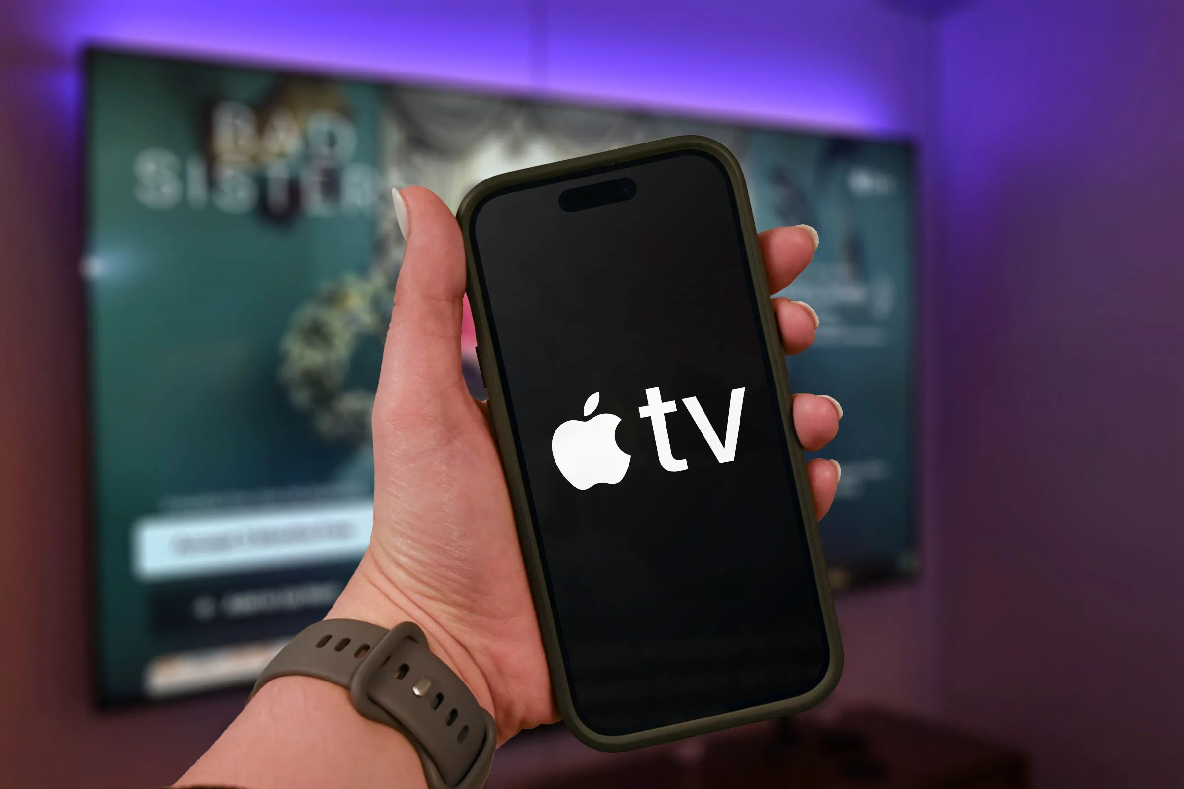Một bàn tay đang cầm iPhone hiển thị logo Apple TV+, với TV mờ ở hậu cảnh.