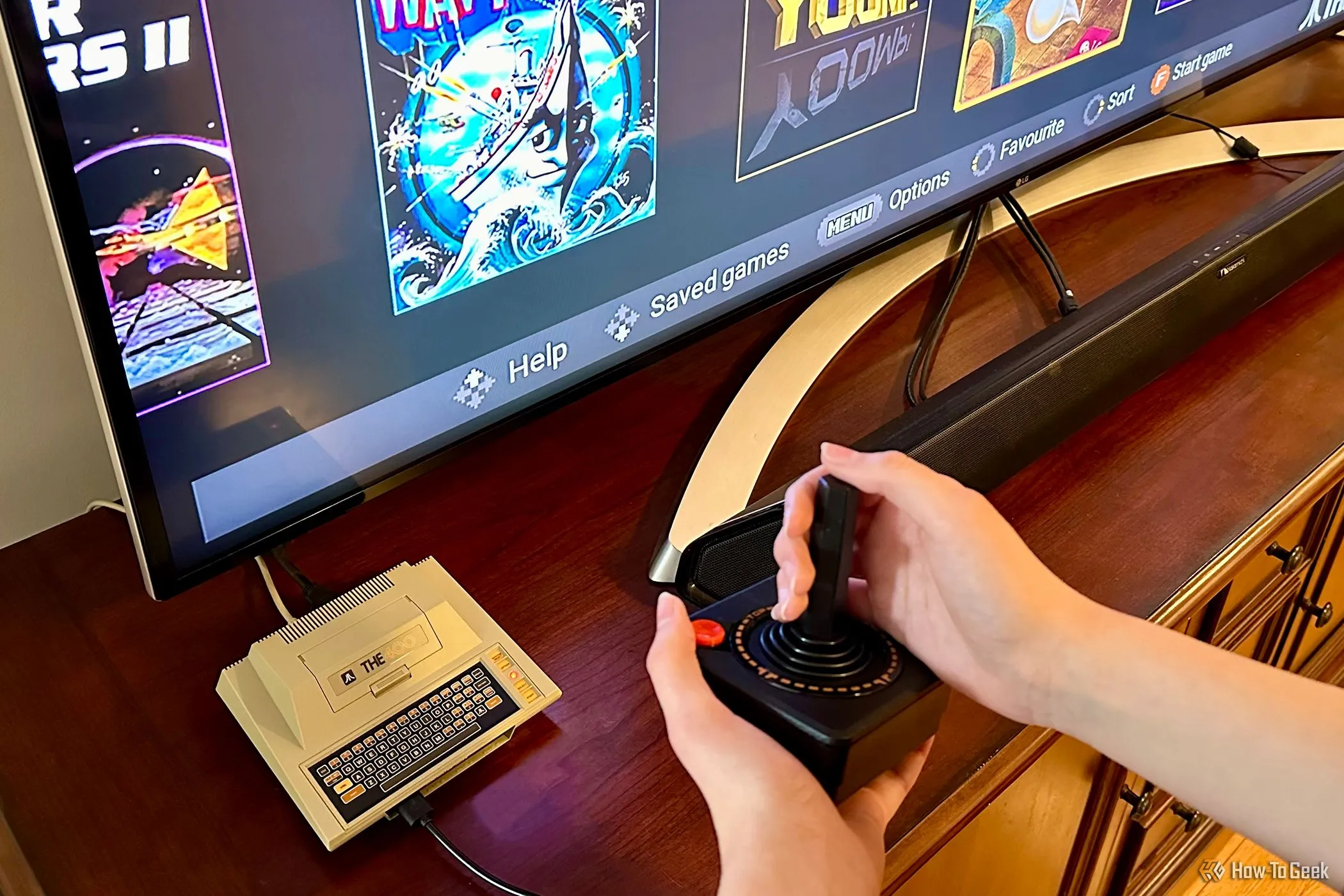 Thiết bị THE400 Mini cạnh TV với đôi tay đang cầm cần điều khiển, gợi nhớ game thùng cổ điển.