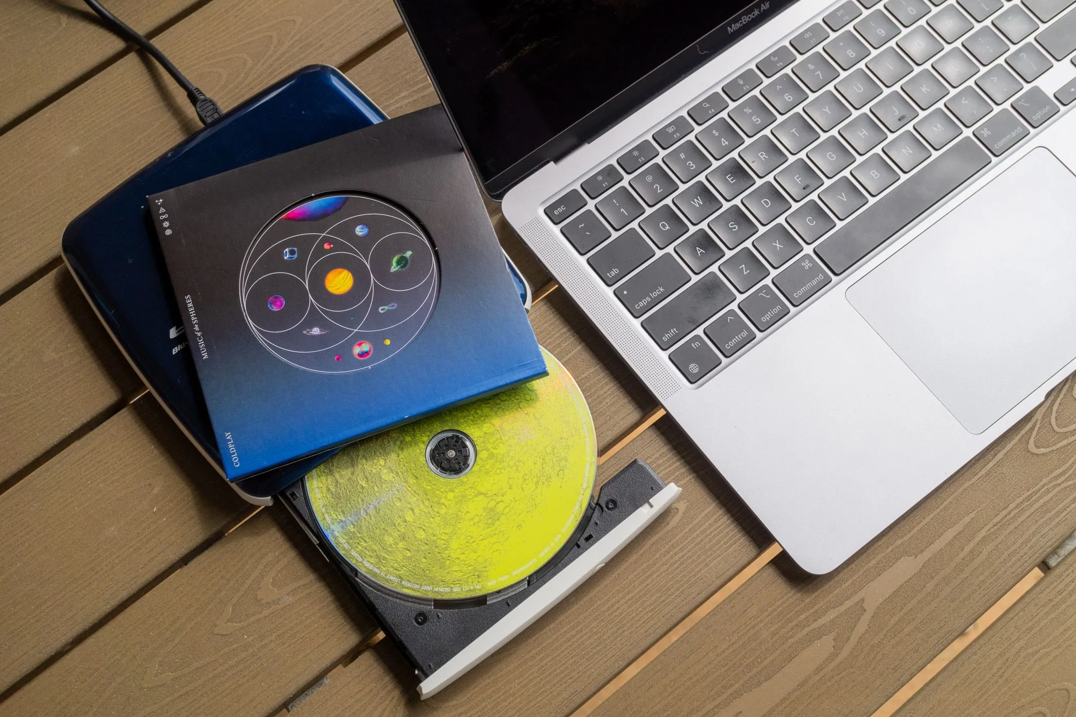 Ổ đĩa CD ngoài và một đĩa nhạc vật lý đặt cạnh MacBook Air, minh họa cách chuyển nhạc từ CD